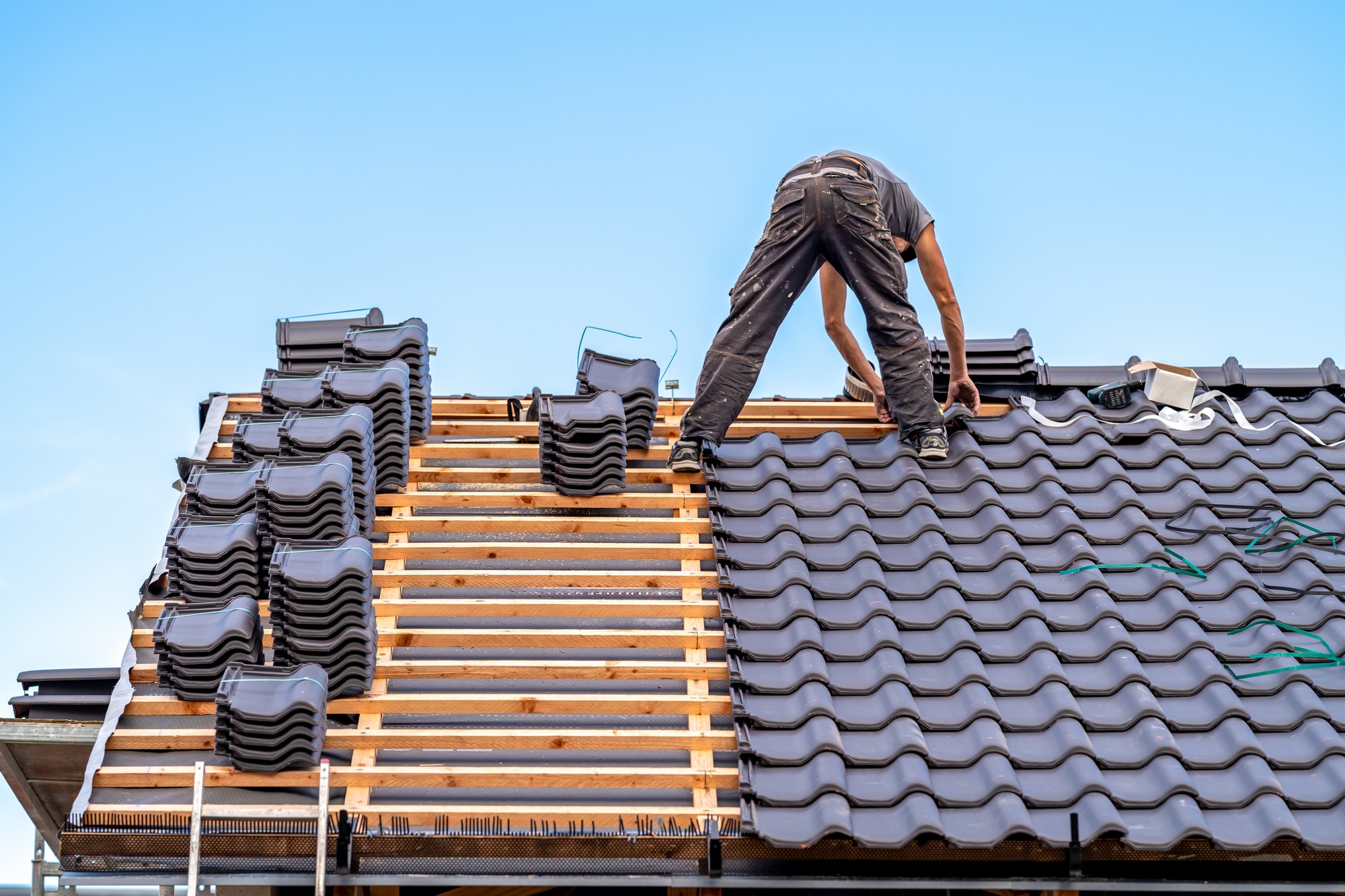 The image shows a person who is installing or repairing a tiled roof. The person is bending over and appears to be adjusting or placing a tile. They are wearing work clothes and are standing on the battens that provide the structure for tile placement. There are stacks of roofing tiles on the roof that have not yet been laid down. The roof installation looks to be in progress, with some areas where the wooden battens are still visible and other areas where the black tiles have been installed. The sky in the background is clear, suggesting nice weather for outdoor construction work. The person is practicing safety by not standing directly on the tiles and seems to be focused on the task at hand.