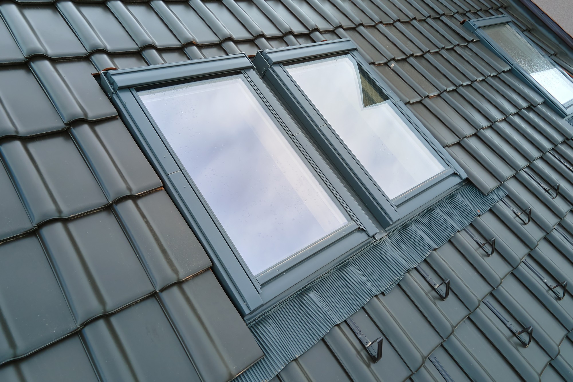 The image shows two windows installed on a tiled roof. The tiles are a dark colour, likely a form of metal or slate roofing, and the window frames appear to be made of a metal or plastic composite, also in a dark colour. There is a flashing used around the windows to ensure waterproofing where the windows penetrate the roof. The sky reflected in the windows suggests an overcast day, and there are a few water droplets on the windows, possibly from recent rain.