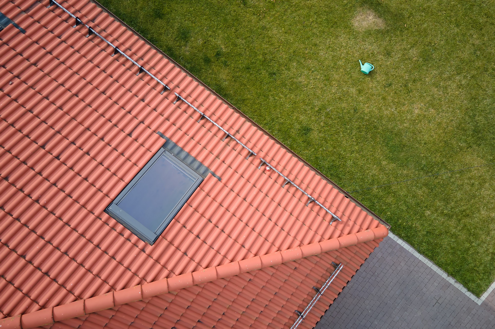 This image shows a top-down view of a building with a sloping terracotta-tiled roof. On the roof, there's a skylight, some metal fixtures, possibly for drainage or anchoring, positioned in a symmetrical pattern. Adjacent to the roof, there's a well-manicured green lawn with a single blue watering can resting on the grass. Alongside the lawn, there appears to be a paved area with grey paving stones or tiles. The vantage point of the image suggests it was taken from an elevated position, looking directly down on the scenes below.