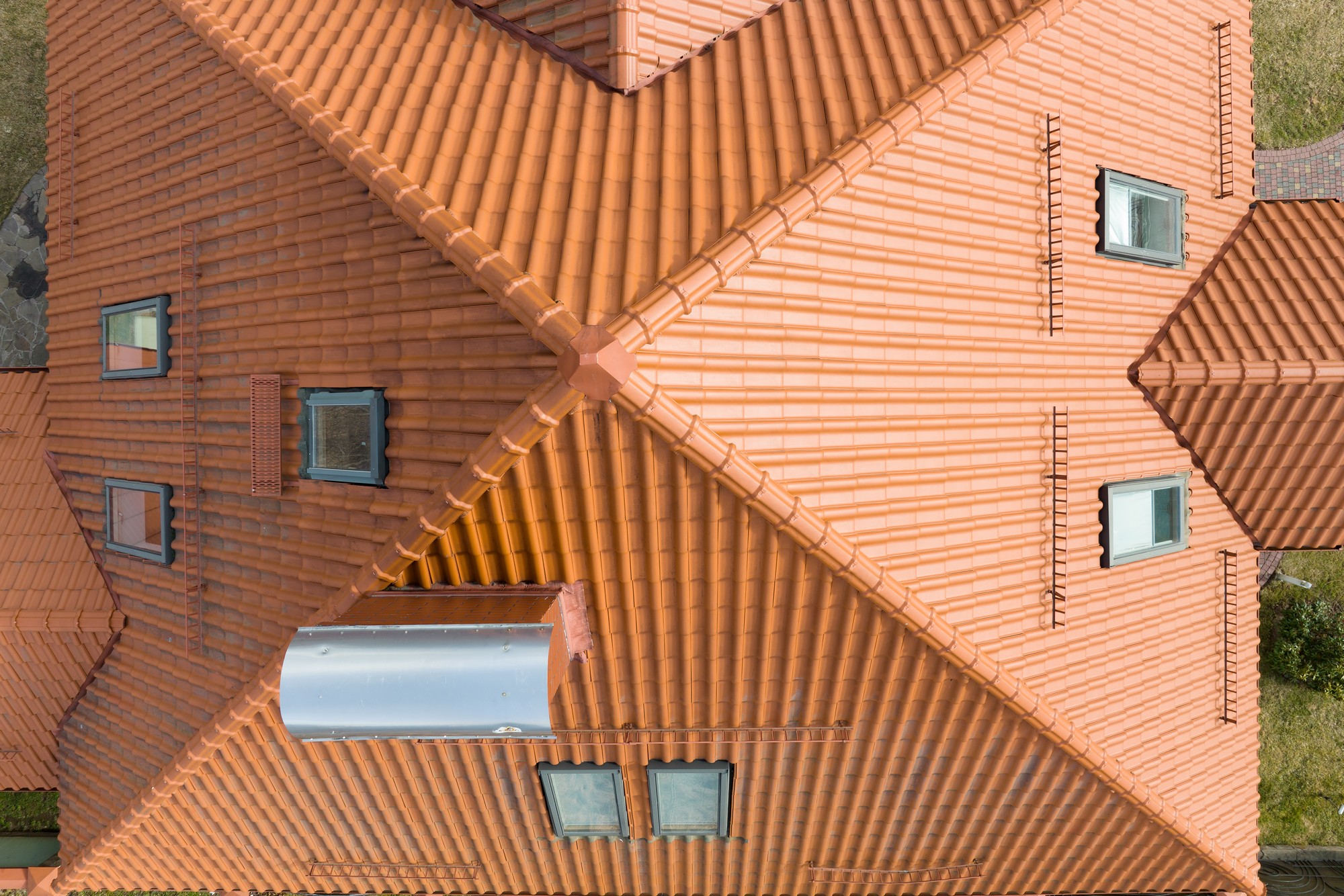 This image shows an aerial view of a building with an orange tiled roof. The pattern of the roof tiles is quite distinctive, with sections of the roof intersecting at various angles, creating a geometric design. There are several dormer windows protruding from the sloped surfaces, each with a pair of windows. There is also a metallic chimney flue or vent pipe on one section of the roof. The gutter system can also be seen along the roof's edges, which collects and directs rainwater away from the building. The surrounding area has sections of green lawn visible at the corners of the image, suggesting the building is in a residential area or has green space around it.