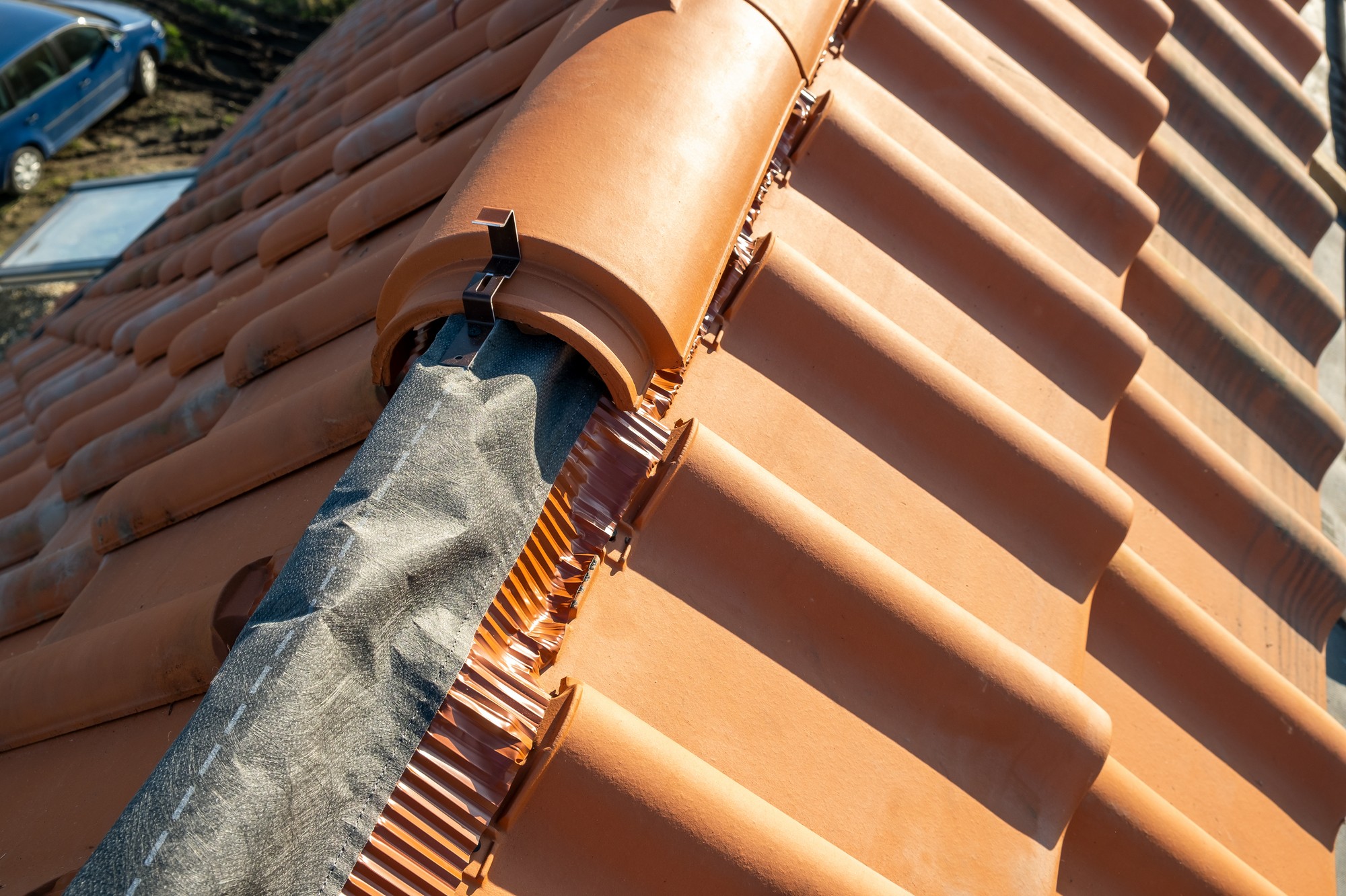 This image shows a close-up of a terra cotta-coloured tiled roof. A roofing ridge is visible, where the two sloping sides of the roof meet at the top. Along the ridge, there is a special ridge tile cap that helps to cover and protect the seam where the two sides join. There's also a weatherproof membrane below the ridge cap, likely for additional protection from the elements. The mounting hardware for the ridge cap is visible, which often includes clips or brackets to hold the cap securely in place. The sunlight indicates it's day, and the shadows indicate it may be early morning or late afternoon. There's also a bit of blue sky in the background and you can see the top of a car at the bottom, indicating that the photo is taken from a higher elevation, such as from the roof itself or scaffolding.