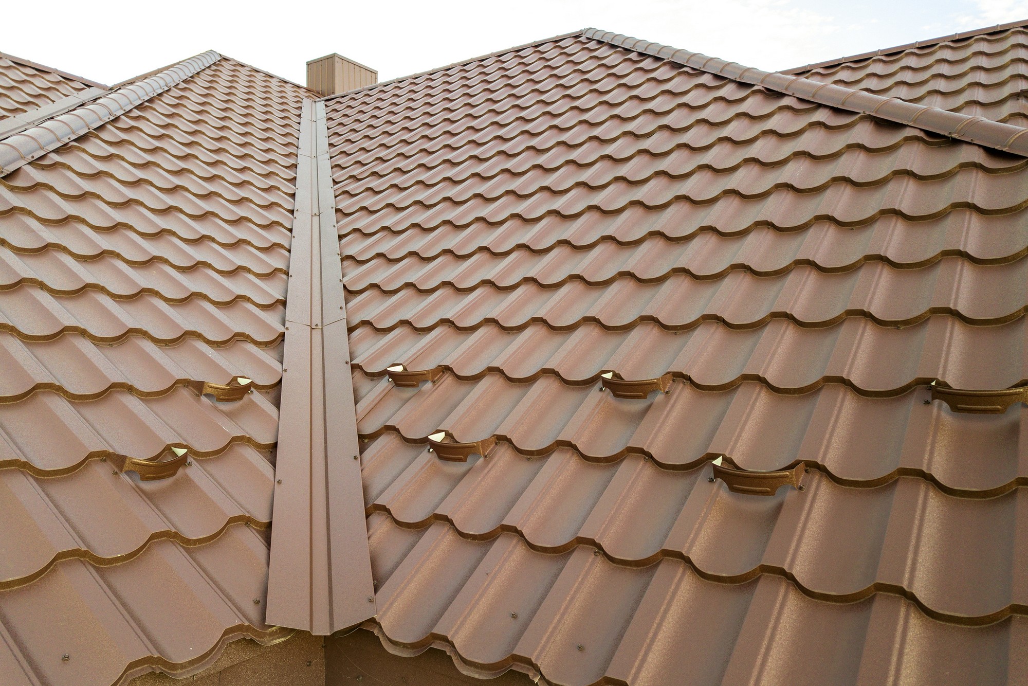 The image displays a close-up view of a terracotta-coloured metal roof. The roof comprises several rows of interlocking metal tiles that mimic the look of traditional clay roofing tiles. You can see the undulating pattern typical for this style of roofing material, which helps with water runoff. There are also visible snow guards installed on the tiles, which are devices used to prevent snow from sliding off the roof in large, dangerous sheets. The snow guards are evenly spaced and attached directly onto the tiles. To the upper left, there appears to be a roof vent, which is likely used for attic ventilation. The perspective suggests that the photo was taken from an elevated position or a level adjacent to the roofline.