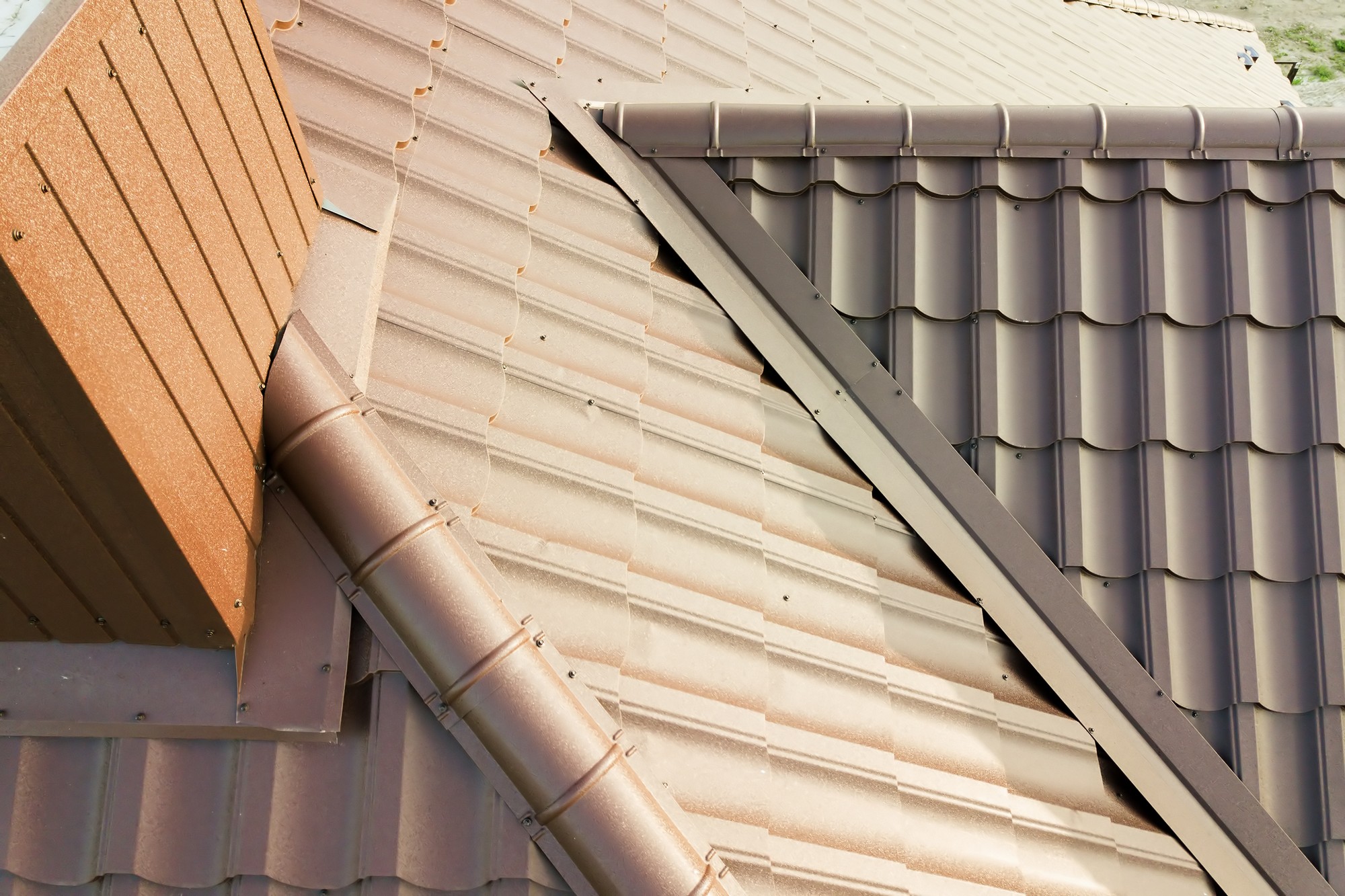 This image shows a close-up of a building's roof. The roof is covered with terracotta-coloured metal tiles on the left side and dark gray metal tiles on the right side. There is a rain gutter system installed on the roof as well, with a downspout visible in the terracotta section. The image also shows a ridge cap and some flashing, which are essential parts of a roofing system used to prevent water from entering the building through joints or gaps. The photo seems to have been taken on a sunny day, given the bright lighting and shadows visible on the roof.