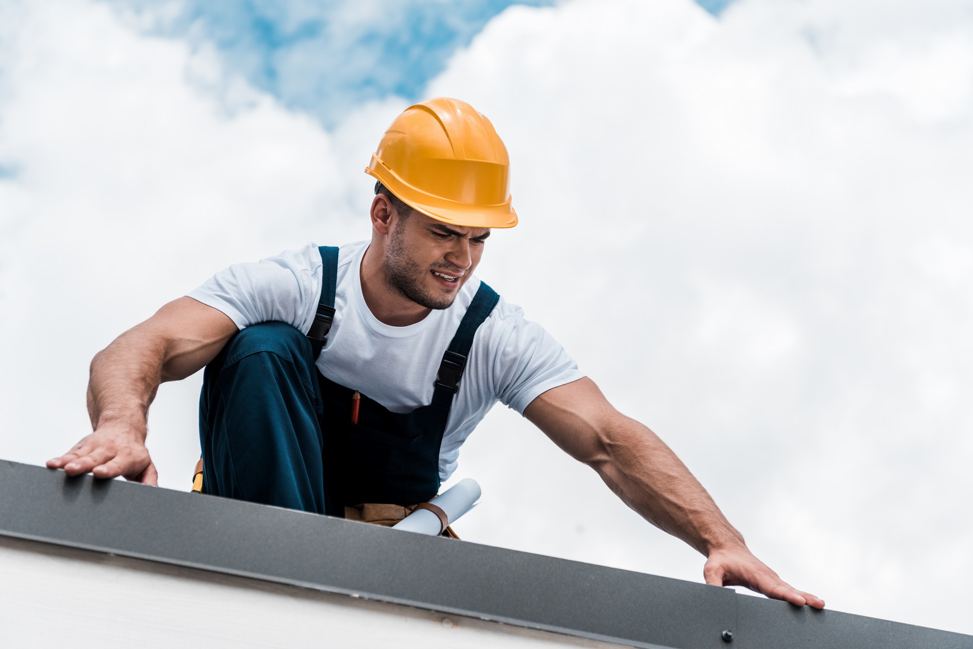 This image shows a person who appears to be a construction worker or contractor. He is wearing a hard hat, which is typically used as protective headgear on construction sites, and is dressed in a white shirt with overalls. The worker also seems to be equipped with a tool belt, suggesting that he may be engaged in some sort of manual work or construction activity. He is reaching over what looks like the edge of a roof or platform, and his expression seems focused, possibly indicating that he is looking at something specific or concentrating on the task at hand. The backdrop is a cloudy sky, which gives the impression that the person is working at an elevated height.