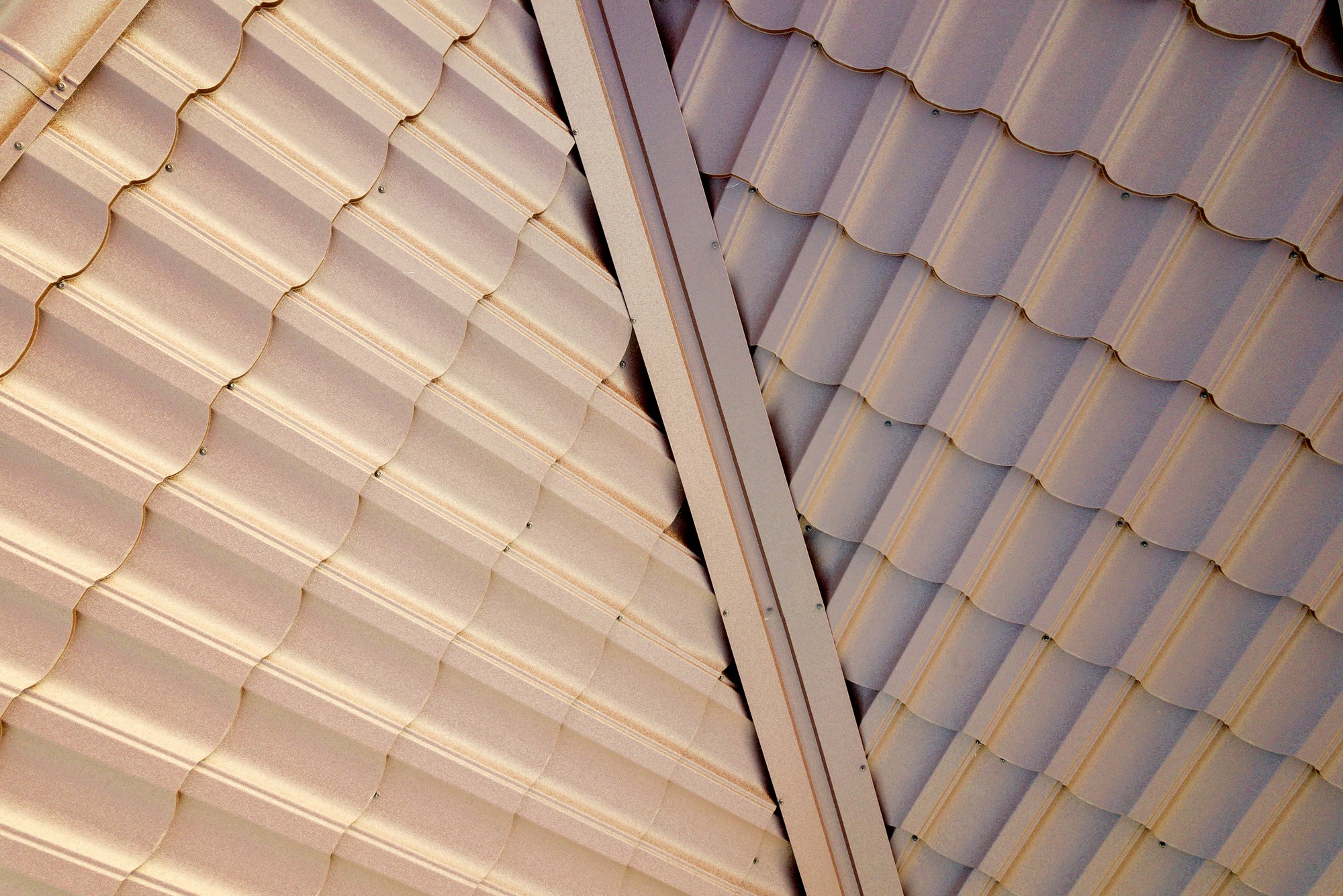 This image shows two sections of a corrugated metal roof meeting at an angle, with one section continuing beyond the other. The corrugations create a wave-like pattern, and the panels are secured with visible fasteners. The lighting and shadows accentuate the texture and form of the metal sheets, while the beige colour lends a warm tone to the image. The angle at which the two sections meet creates a visually interesting diagonal line across the frame.