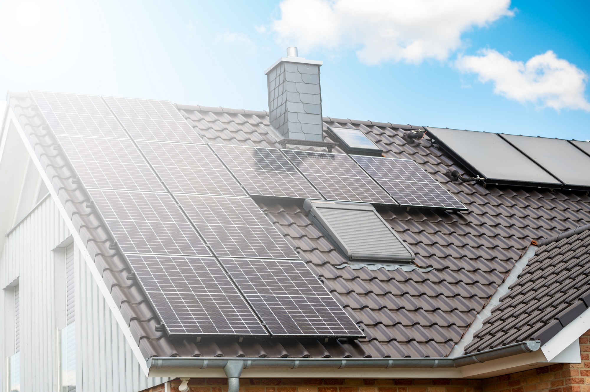 The image displays a residential roof equipped with several solar panels. These panels are installed to harness solar energy, converting it into electricity to power the home. The roof appears to have a sloped design with dark-coloured tiles. There's also a chimney visible on the roof, and a part of the house's structure shows on the left side of the image. The bright sunlight and blue sky suggest that the conditions are favorable for solar energy generation.