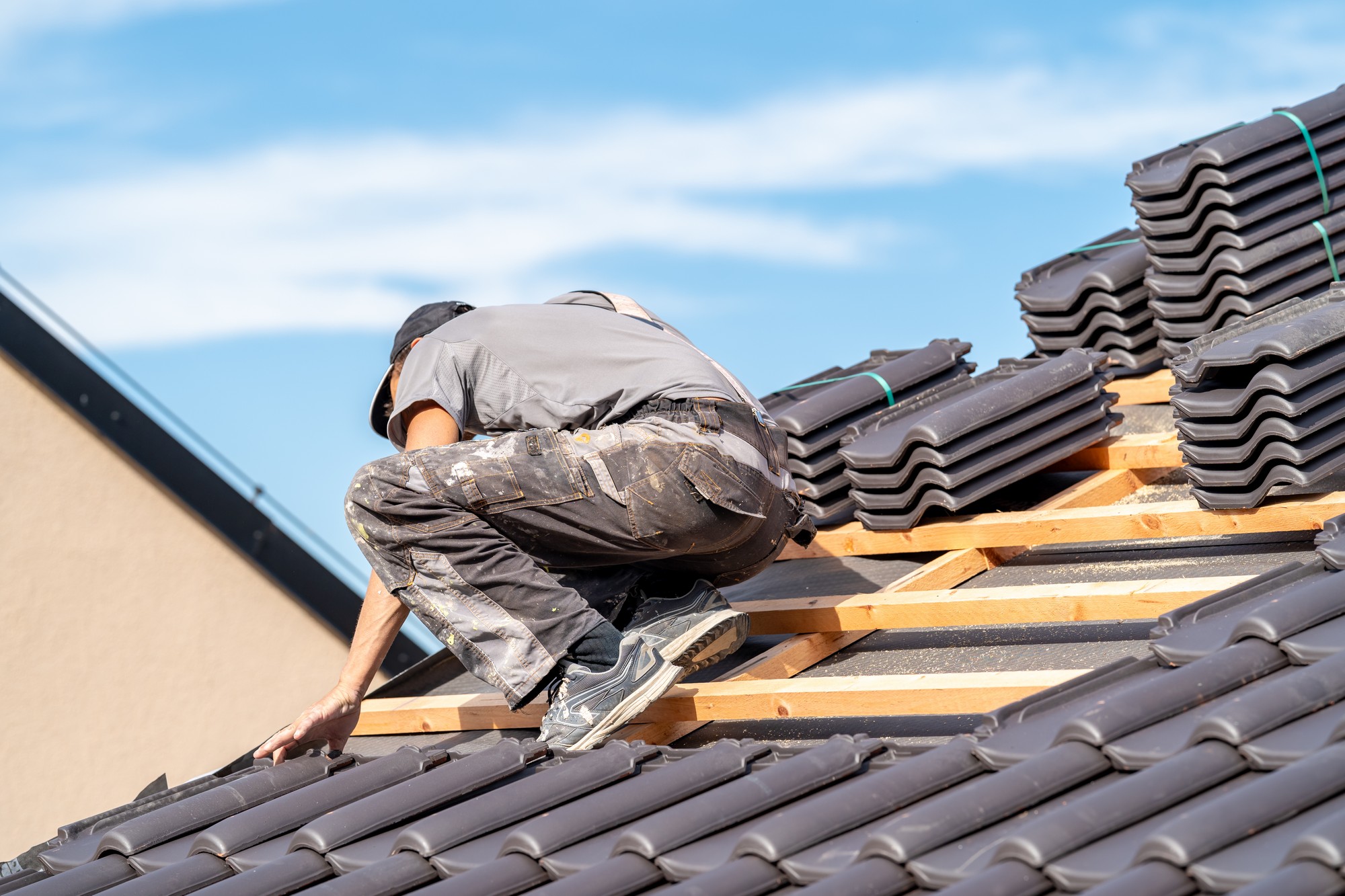 The image displays a person engaged in the process of roof installation or repair. They are crouched down on a sloped roof structure that is partly covered with roofing tiles. The person is wearing a baseball cap, a short-sleeve shirt, and pants that are marked with what appears to be paint or roofing materials, indicating they're actively working. In the foreground, you can see a stack of unused roofing tiles, suggesting the roofing job is still in progress. The person is wearing gloves for safety, and the clear blue sky suggests that the work is being conducted on a day with favorable weather conditions. The framing is diagonal, emphasizing the slant of the roof and creating a dynamic composition.