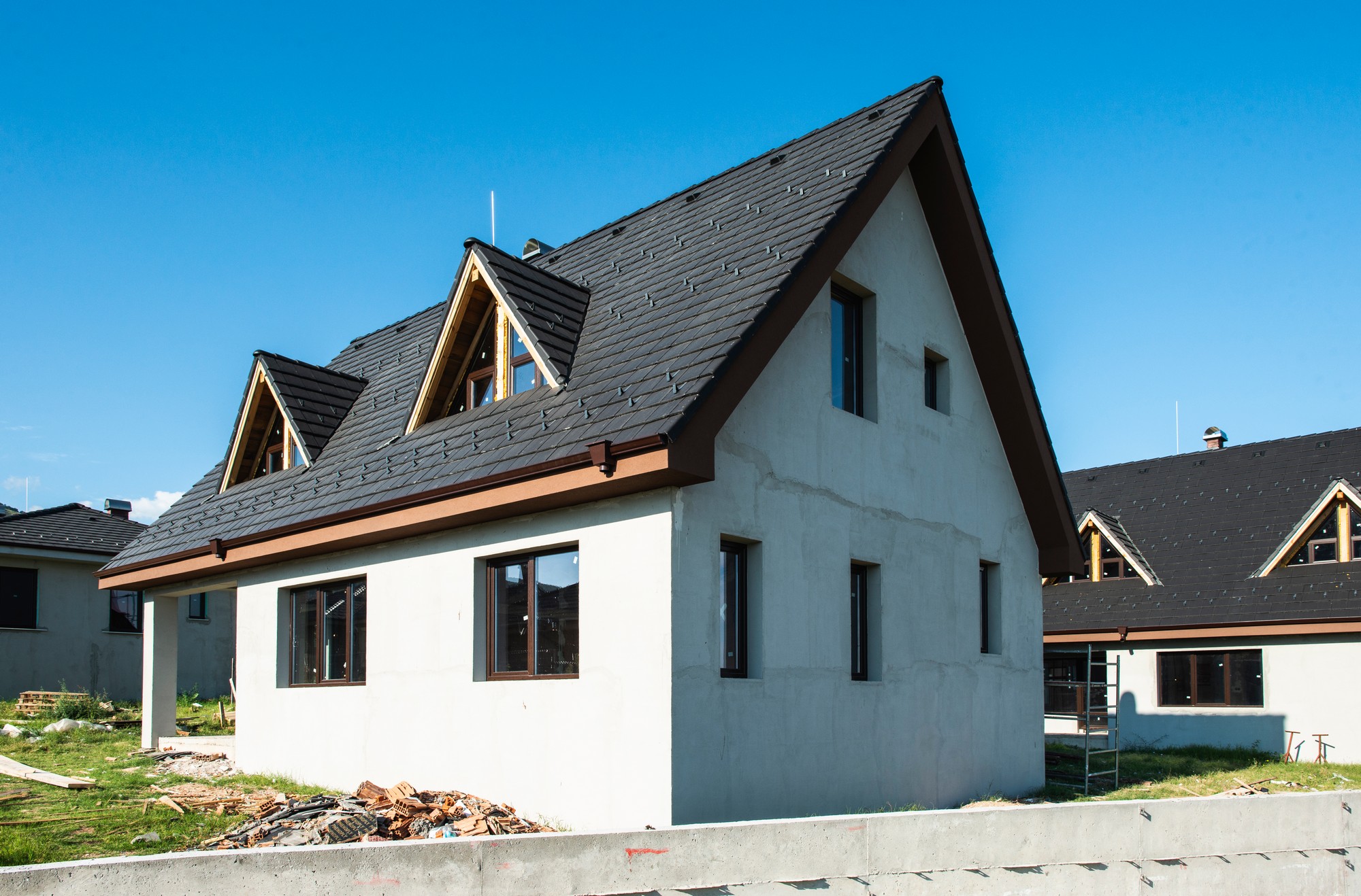 The image shows a house that appears to be under construction or recently constructed. The house has a pitched roof with dark shingles, dormer windows, and a combination of finished areas with siding and areas with exposed construction materials. The exterior walls are partially covered with what looks like a white plaster or stucco finish. There are window openings, some of which are already fitted with windows, while the others are merely openings. Debris and construction materials can be seen around the house, indicating ongoing work. The ground is not landscaped and looks like a typical construction site with a mix of dirt and grass. The skies are clear and blue, suggesting good weather conditions.