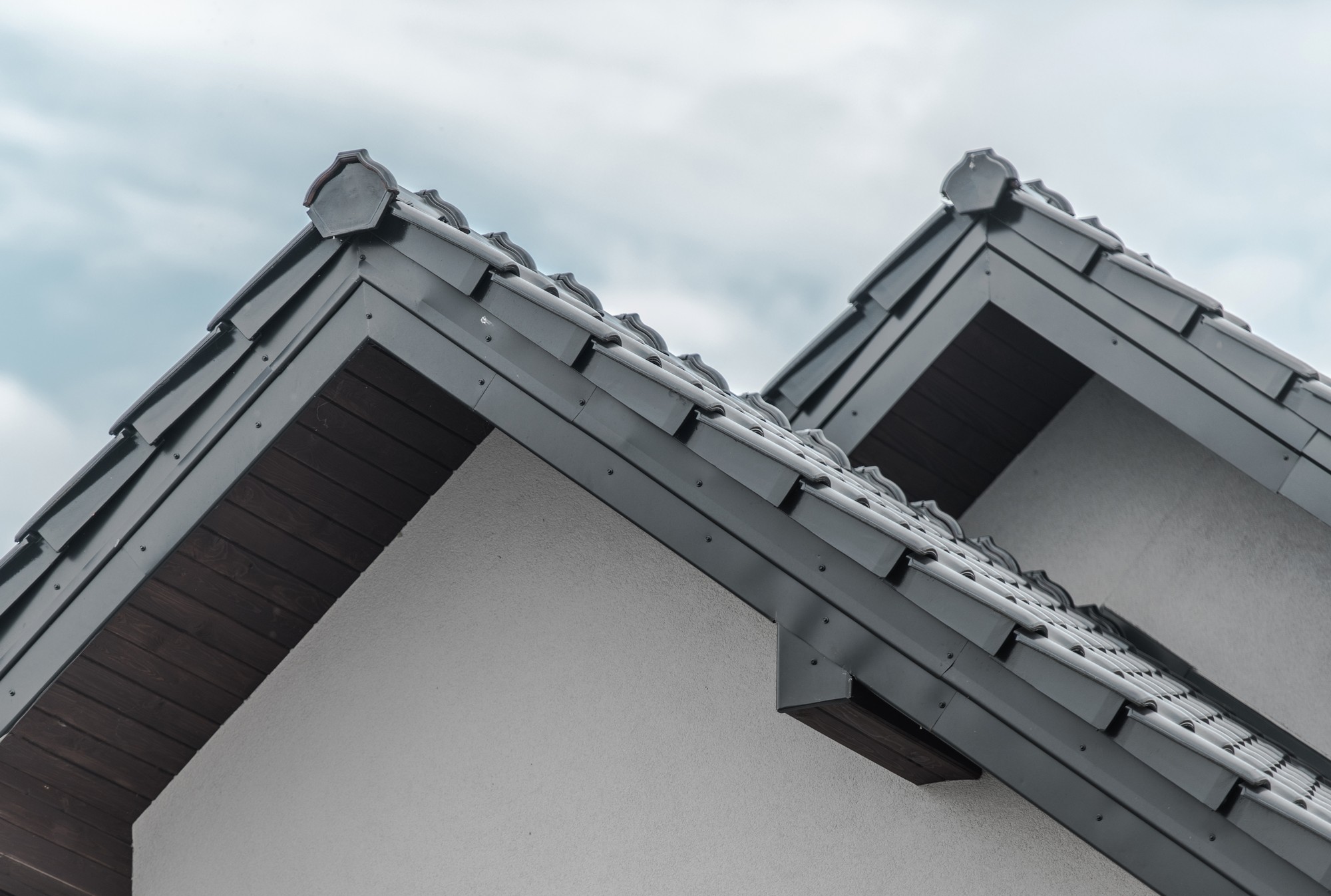 The image shows the upper part of a building focusing on its roof details. Specifically, it features two sloped sections of a tiled roof with ridge caps at the apex where the two slopes meet. The roof tiles appear to be modern, possibly concrete or clay tiles. You can also see the roof's overhang with wooden soffits or eave boards under the eaves, adding detail and aesthetics to the design. The gutters or rainwater collection systems are not visible in this particular shot. The colours are neutral, with the roof tiles in a dark grey, the ridge caps in a matching colour, and the soffits in a dark brown, which contrasts nicely against the light-coloured stucco or rendered finish on the walls. The sky in the background is overcast, suggesting that the weather is cloudy.