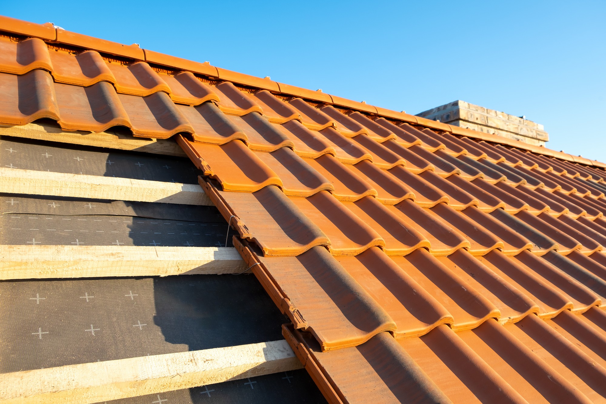 The image shows a close-up view of a terracotta tiled roof. The roof tiles are laid in an overlapping pattern, typical for clay or ceramic roofing materials, designed to shed water and protect the structure from the elements. You can see the wooden roof battens to which the tiles are secured, and beneath that is a layer of roofing underlayment, which provides additional water protection. The angle of the photo and the lighting suggest it may be taken during the daytime with a clear sky.