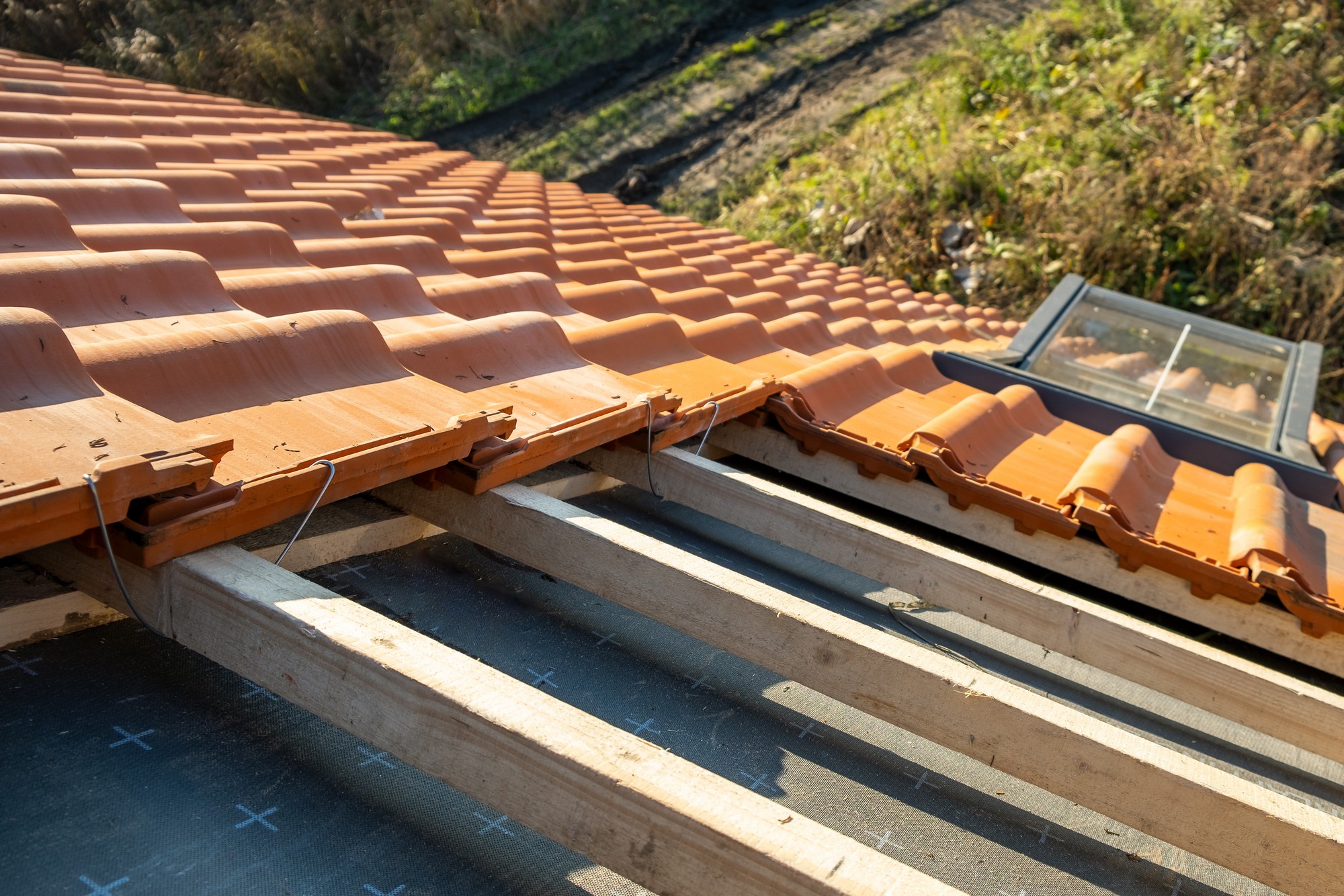 The image shows a close-up view of a terracotta-coloured tiled roof under construction. You can see the wooden roof beams and the black waterproof sheeting between them, which is typically used to provide an additional layer of protection against moisture. Some of the roof tiles are already in place, while others are not, revealing the underlying structure. There's also a roof window, sometimes called a skylight, installed on the roof, which is designed to bring natural light into the space below. The photo captures the details of the materials and construction methods used in roofing.