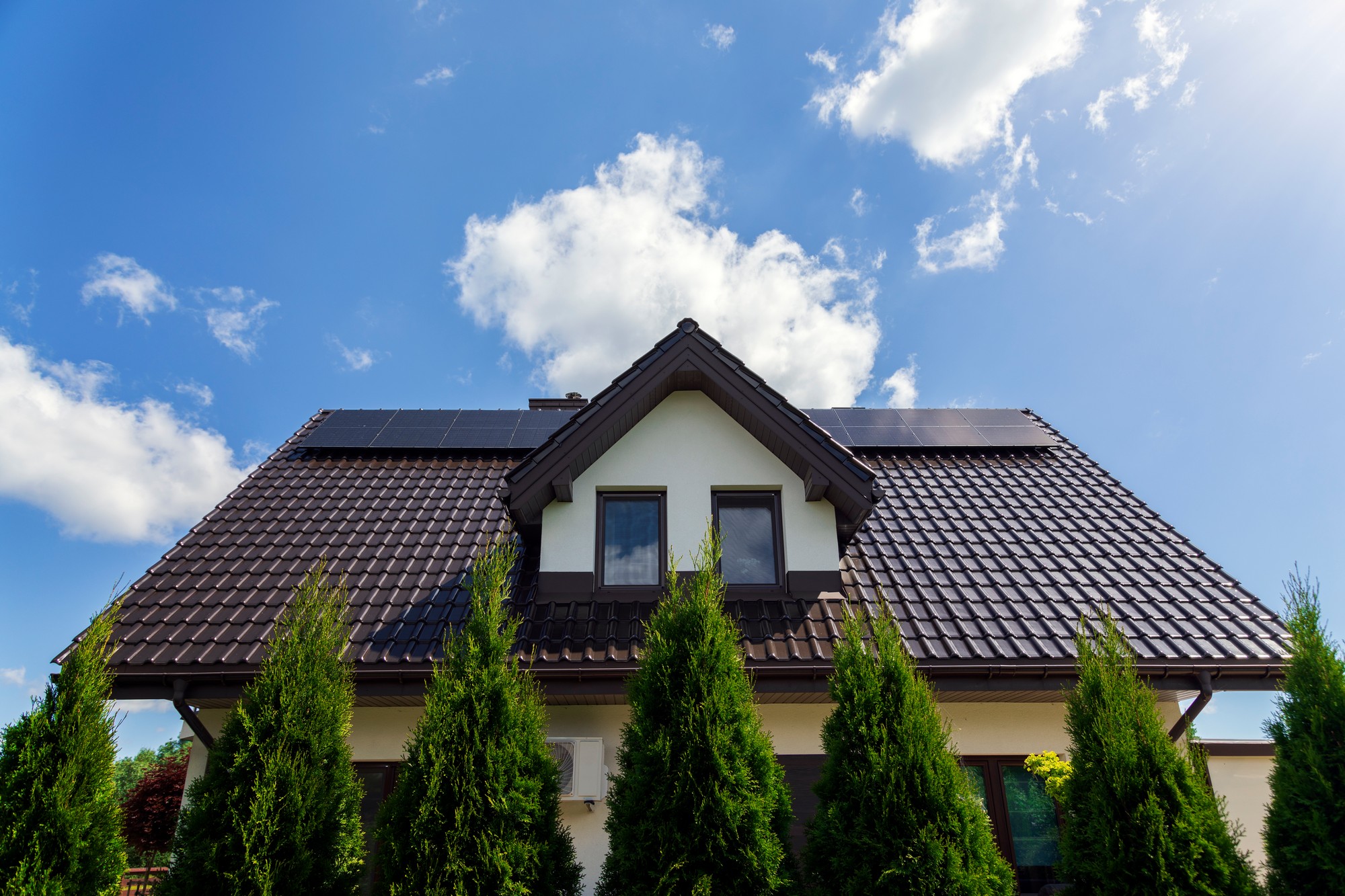 This image shows the upper part of a house with a pitched roof that has solar panels installed on it. The house appears to be two-storied, with visible windows on the upper level. Around the building, there are several tall, narrow coniferous trees, possibly Thuja, planted closely together which might be serving as a hedge or ornamental feature. The sky is blue with a few scattered clouds, and there's a glimpse of sunlight that suggests a clear, sunny day. The installation of solar panels indicates a use of renewable energy sources for electricity generation for the home.