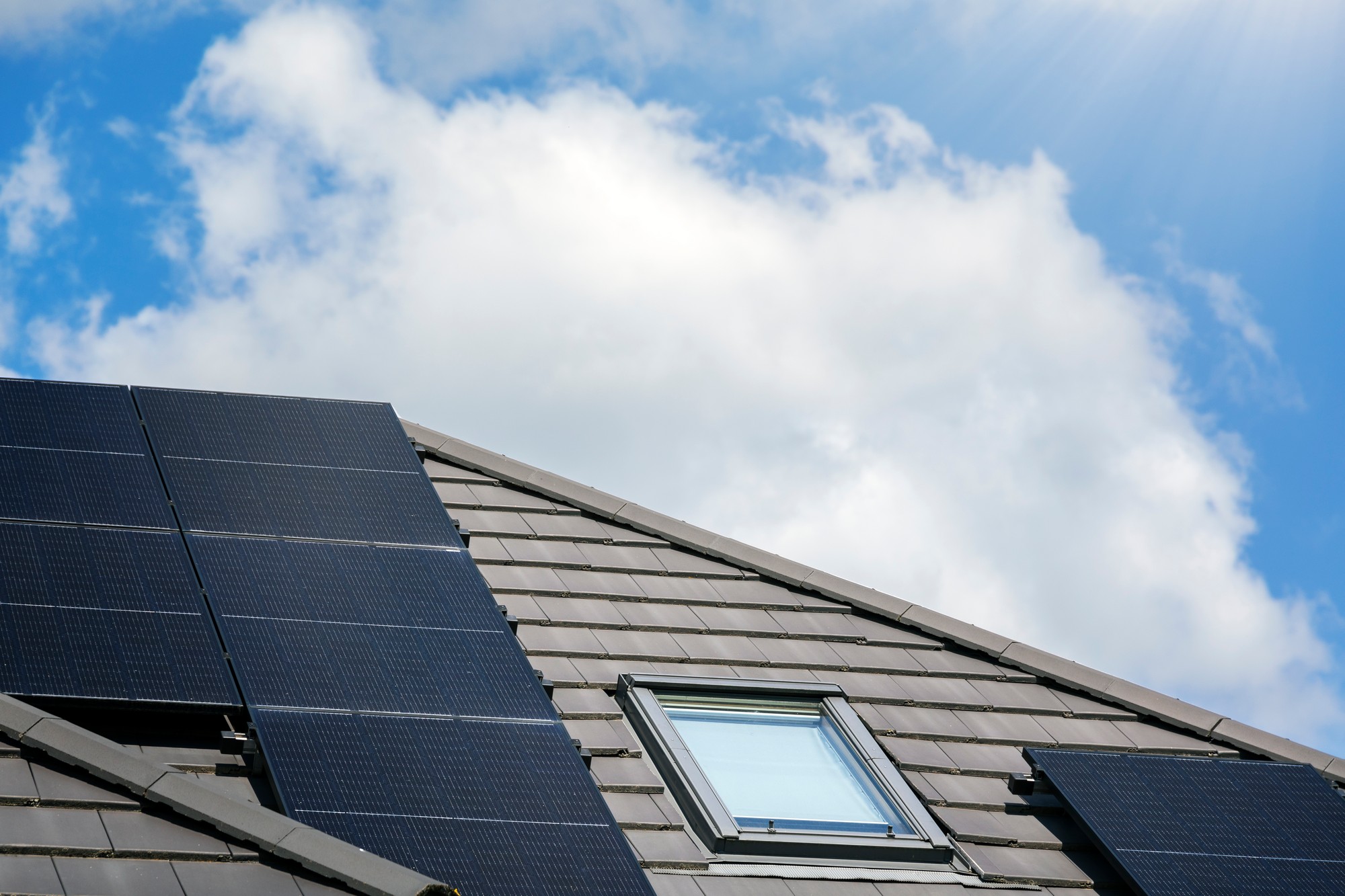 The image shows a section of a sloped roof with solar panels installed on it. There is also a skylight window integrated into the roof. The blue sky with some clouds is visible in the background, and sun rays are peeking from the upper right corner, suggesting a sunny day which is ideal for solar energy production. These solar panels capture sunlight and convert it into electricity, contributing to a home's energy supply and reducing reliance on traditional energy sources. The presence of the skylight indicates an interest in utilising natural light within the building as well.