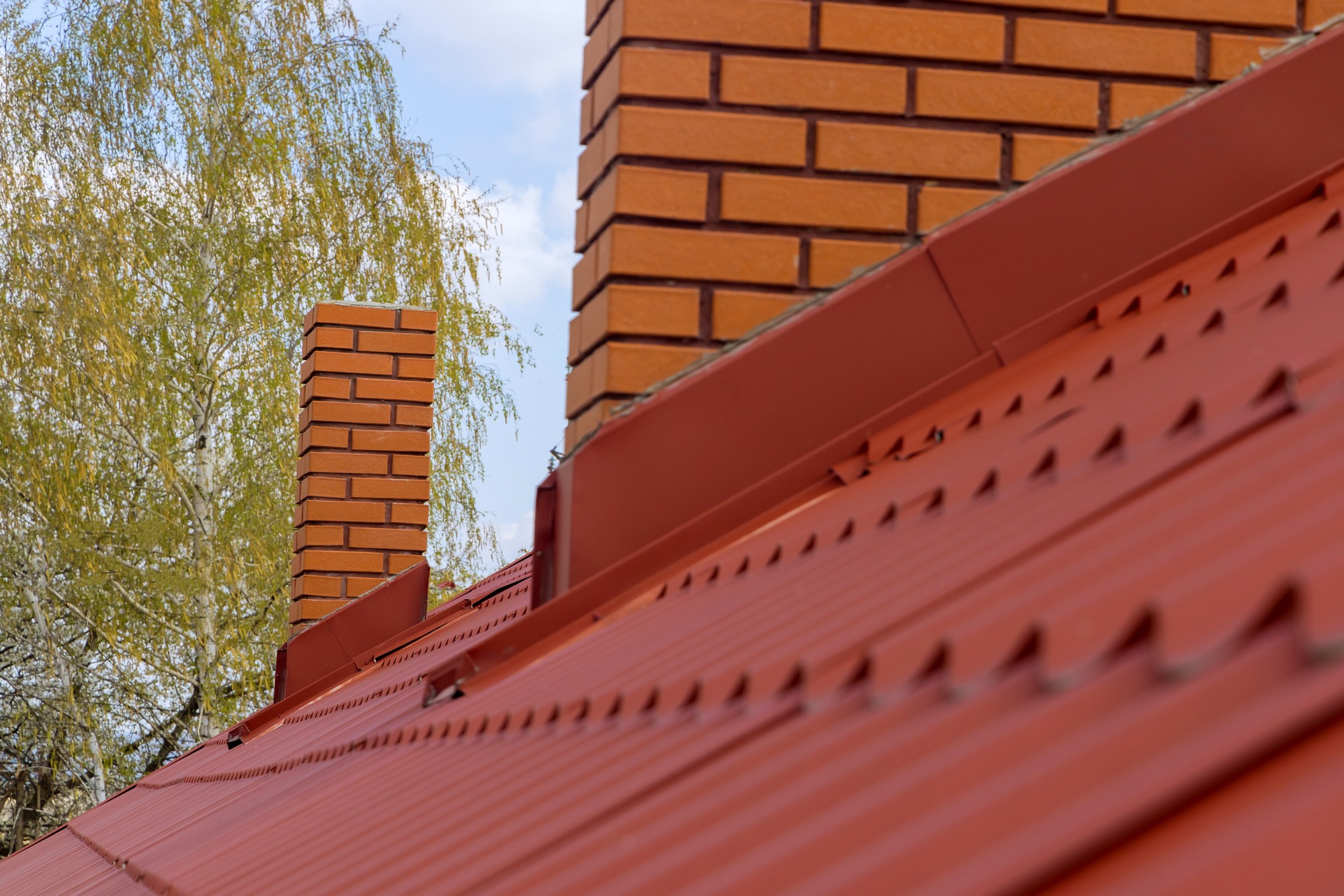 The image depicts a section of a building with a focus on the architectural details. In the foreground, there's a metal roof with a distinctive red hue, showing the corrugated pattern and fastening screws. Slightly out of the main focus, in the midground, we see the top part of a brick chimney extending above the roofline. The bricks also have a similar red colour which complements the metal roof. In the background, beyond the roof and chimney, there's a blurred view of a tree with green leaves, suggesting an outdoor setting and likely a residential area. The sky appears to be partly cloudy, allowing for a soft, natural light in the scene.