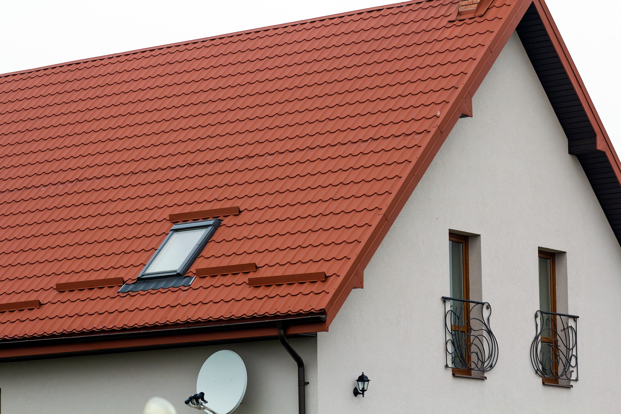 The image shows a part of a residential building, primarily focusing on the sloped roof covered with red tiles. A roof window, also known as a skylight, is integrated into the roof structure, allowing natural light into the attic or room below. There are also decorative elements such as window boxes with wrought iron designs on two windows, which are adding a touch of elegance to the facade. The walls are painted in a light colour, which contrasts with the dark-red roof tiles. To the side, there is a satellite dish mounted on the building, suggesting the occupants have satellite television services. A small exterior light fixture is installed near the windows, likely for outdoor illumination. The overall appearance suggests a well-maintained, suburban or rural home.