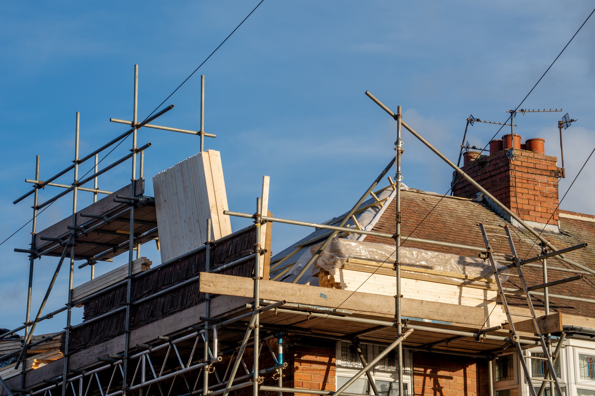 The image shows a residential building undergoing construction or renovation with scaffolding erected around it. The scaffolding is made up of metal poles and wooden boards that provide a platform and support for construction work. There is a blue sky in the background, suggesting it might be a clear day. The roof of the house is partially visible, featuring exposed wooden rafters and plastic sheeting, likely indicating that roof work is being performed. There's also a chimney with multiple flues at the very top, and various electrical or telephone wires crossing in front of the house.