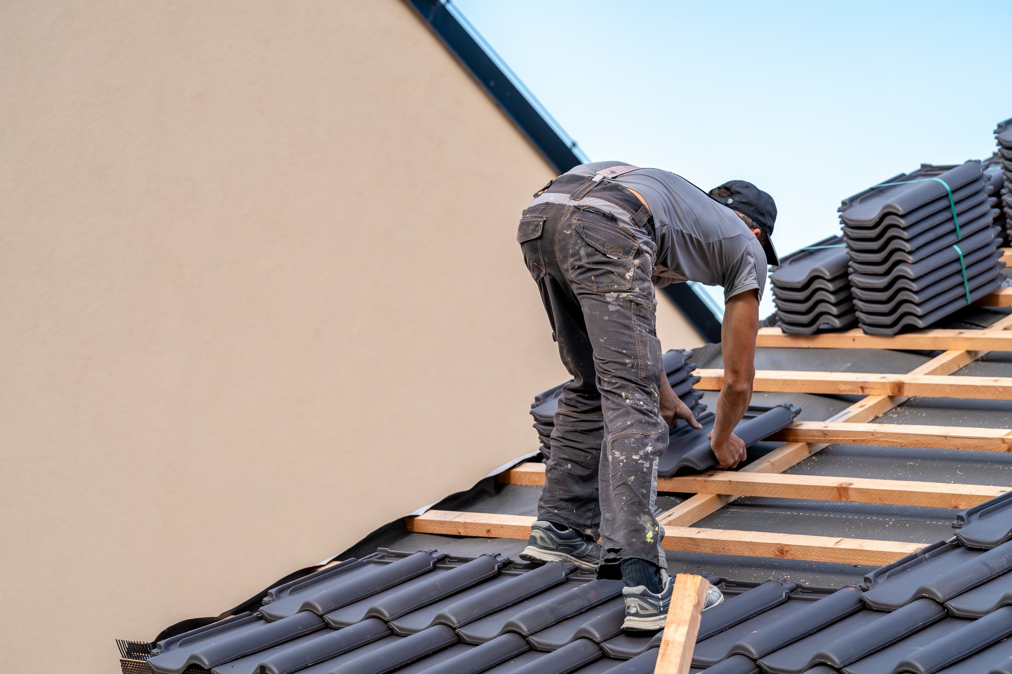 This image features a person working on the roof of a building. They are in the process of installing or repairing roof tiles. The person is bent over, working on the tile alignment, and appears to be wearing work clothing that includes a cap, gloves, and sturdy shoes. The roof structure is partially completed, with wooden battens showing where tiles have not yet been laid.

In the background, we see a neatly stacked pile of additional tiles that are ready to be installed. The sky is clear, suggesting fair weather conditions for outdoor work. The angle and composition of the photo focus on the worker and highlight the hands-on, skilled labour involved in roofing.