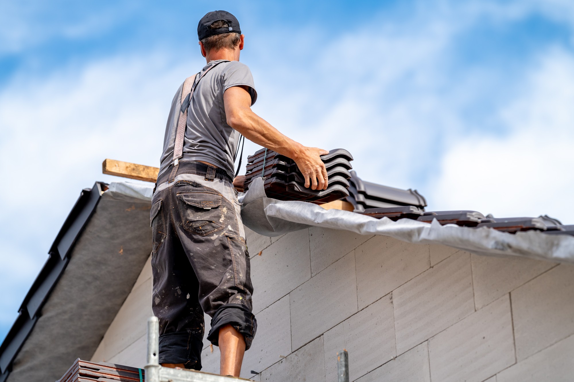 The image shows a person working on a roof. The individual, likely a construction worker or roofer, is seen from the back and appears to be installing roof tiles. They are wearing casual work clothes, including a short-sleeved shirt, trousers with visible pockets, and a cap. The sky behind them is partly cloudy, which suggests it is an open-air environment, typical for roofing work. The worker is handling roofing materials, and there is a protective layer on the part of the roof that's visible, which might be roofing underlayment. Safety measures like the use of scaffolding or a harness are not visible in the image, but these are common safety precautions for such work.