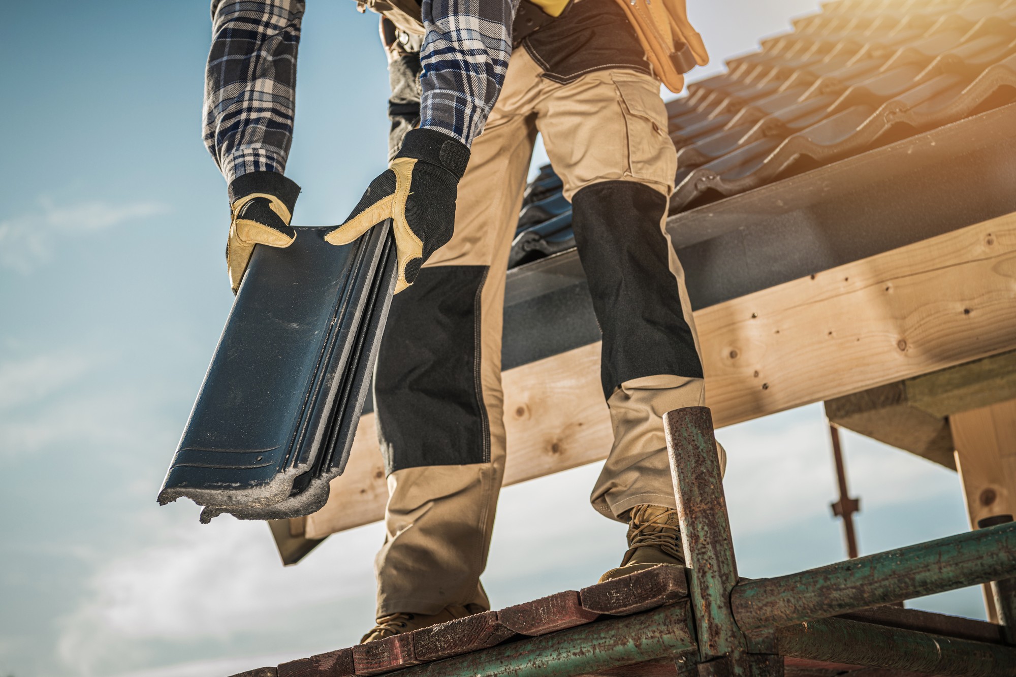 The image depicts a person engaged in roofing work. They are standing on a ladder and appear to be installing or replacing roof tiles or shingles. The individual is wearing protective gloves, a long-sleeve plaid shirt, and work pants, indicating they are appropriately dressed for construction or manual labour. You can also see a part of a wooden structure, which could be the roof they are working on, and the clear sky in the background suggests it might be good weather for outdoor work.