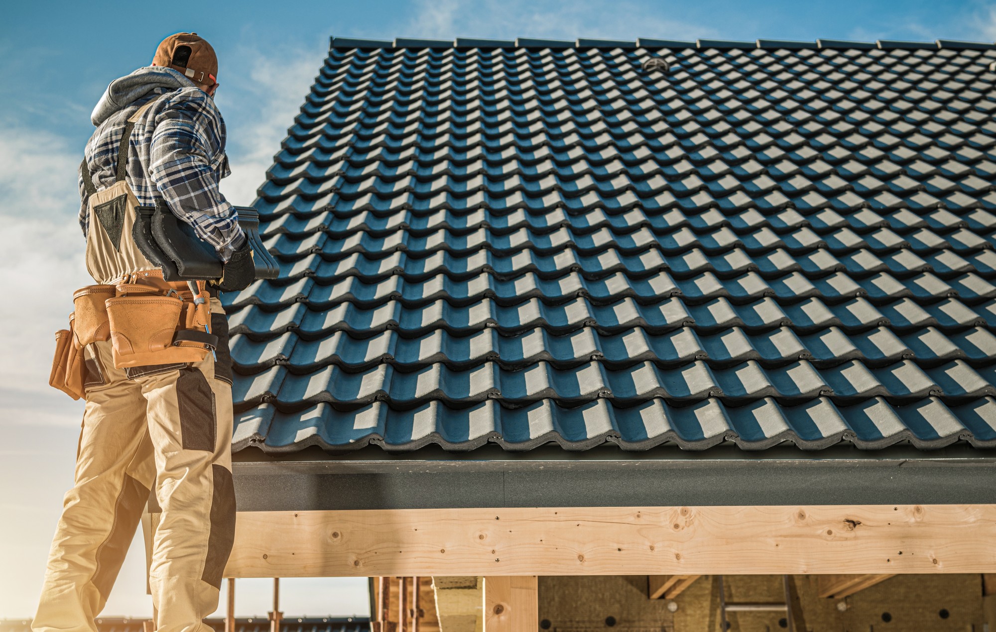 The image shows a person standing on a scaffold, working on a roof fitted with dark-coloured tiles. The individual is dressed in work attire, complete with a checked shirt, a hoodie, work pants, a tool belt, and a hard hat. The roof appears to be new construction or undergoing renovation, with a clean and orderly arrangement of tiles visible. The sky is clear, suggesting favorable weather conditions for outdoor construction work.