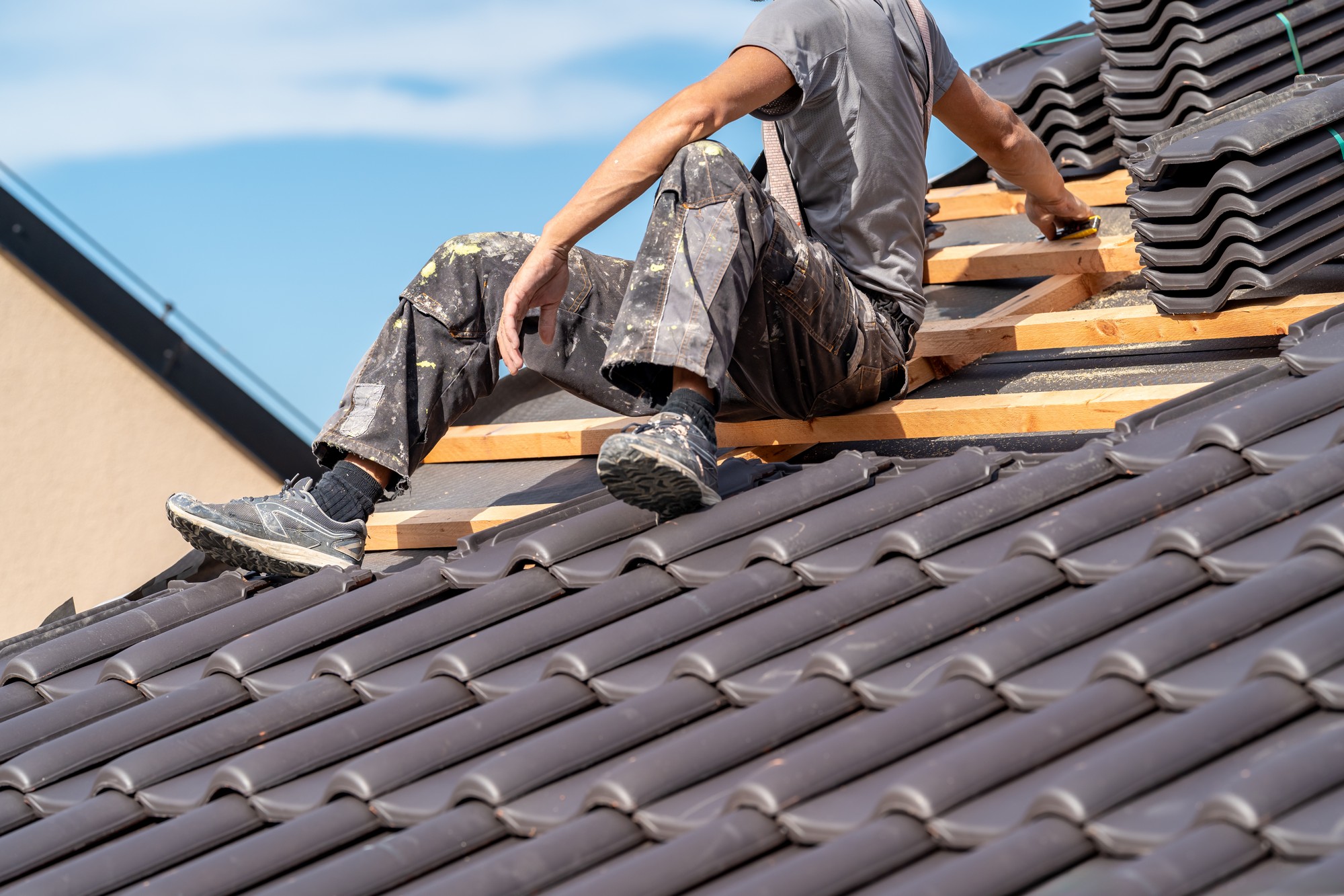 The image shows a person sitting on a roof that is under construction or repair. The person appears to be a worker, as indicated by the work attire that includes a utility belt, pants with paint marks, and sturdy shoes. They are amidst roof tiles that have been partially laid down. The roofer is seen measuring and possibly cutting a wooden board, which is part of the roof structure. Behind the worker, there's a clear blue sky indicating good weather conditions for outdoor construction work.