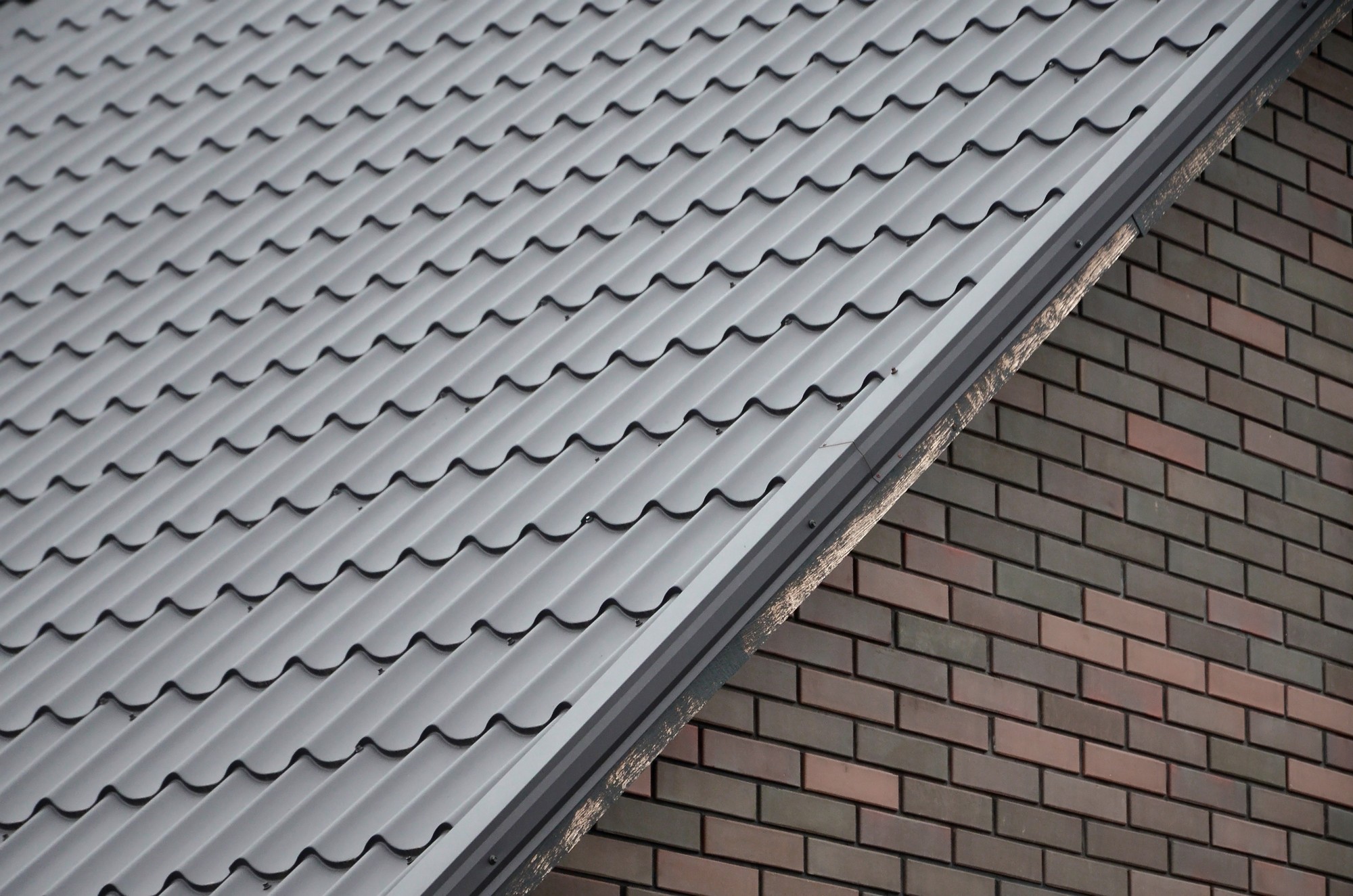 This image displays a close-up of a building, showing two distinct areas: a section of dark gray, corrugated metal roofing on the upper left and a section of brick wall on the lower right. The metal roof exhibits the S-shape pattern typical of corrugated roofing materials, designed to provide strength and water resistance. The brick wall consists of bricks laid in a uniform pattern, with a range of brown and red hues. A thin metal flashing is visible at the intersection of the roof and wall, which is generally used to prevent water from seeping into the building at joints or edges. The overall composition and angle create a visually interesting contrast between the textures and patterns of the roofing and brick materials.