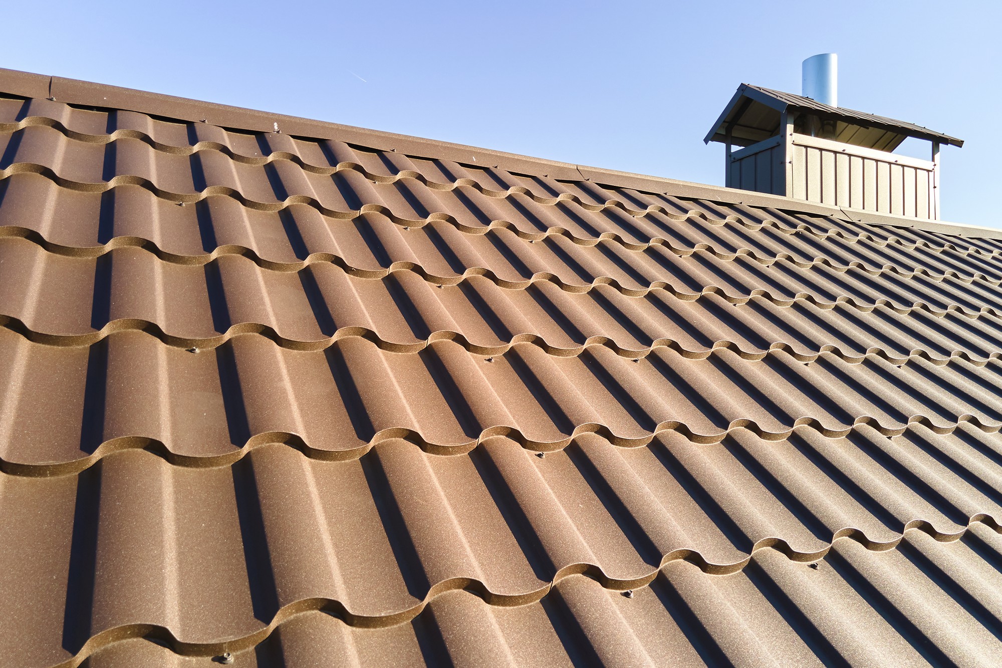 The image shows a close-up view of a corrugated metal roof with a chimney stack visible in the background. The metal sheets have a wavy pattern, which is typical for this kind of roofing material as it provides strength and allows for efficient water runoff. The colour of the roofing appears to be brown or a similar earth tone, and the sky in the backdrop is clear and blue, indicating fair weather. The chimney stack has a protective cap on top, which is often used to prevent rainwater from entering and to keep birds and other animals out.