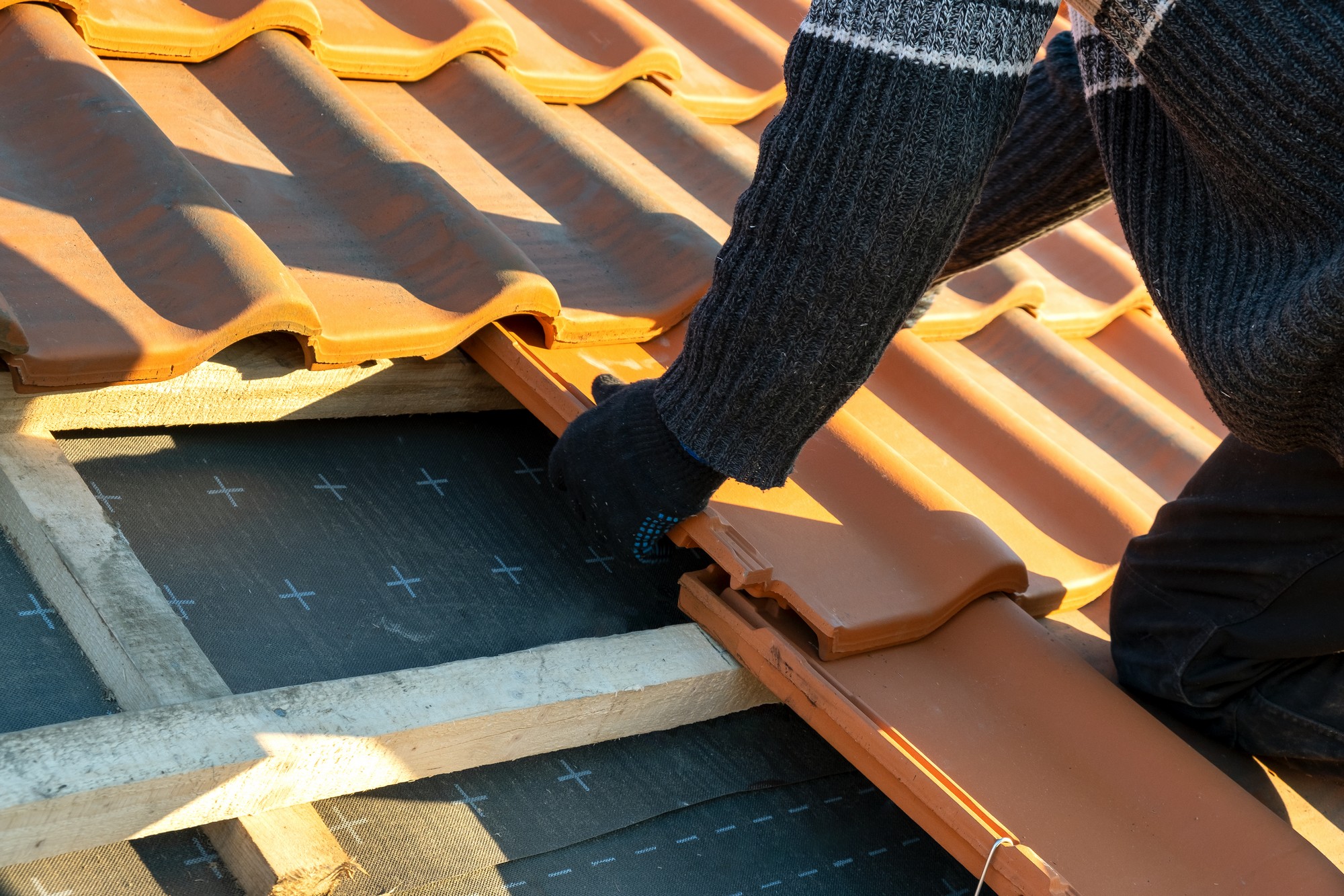 This image shows a person working on a roof. The individual is in the process of laying down terracotta-coloured roofing tiles. To the left, some tiles have already been placed, while in the centre, a section of the wooden roof structure is visible, with what appears to be a waterproof black underlayment material.

The person's hands, which are wearing protective gloves, are adjusting a tile into position. The person is dressed in clothing appropriate for manual labour, including a knit jumper and dark trousers, indicating that the work is being done in an environment that may be cool or requires some level of protection.

The sunlight and shadows on the roof suggest it could be either morning or late afternoon, giving the work environment a bright and clear condition which is often ideal for construction work. Overall, the image captures a moment of roofing construction, with a focus on the tiles and the installation process.