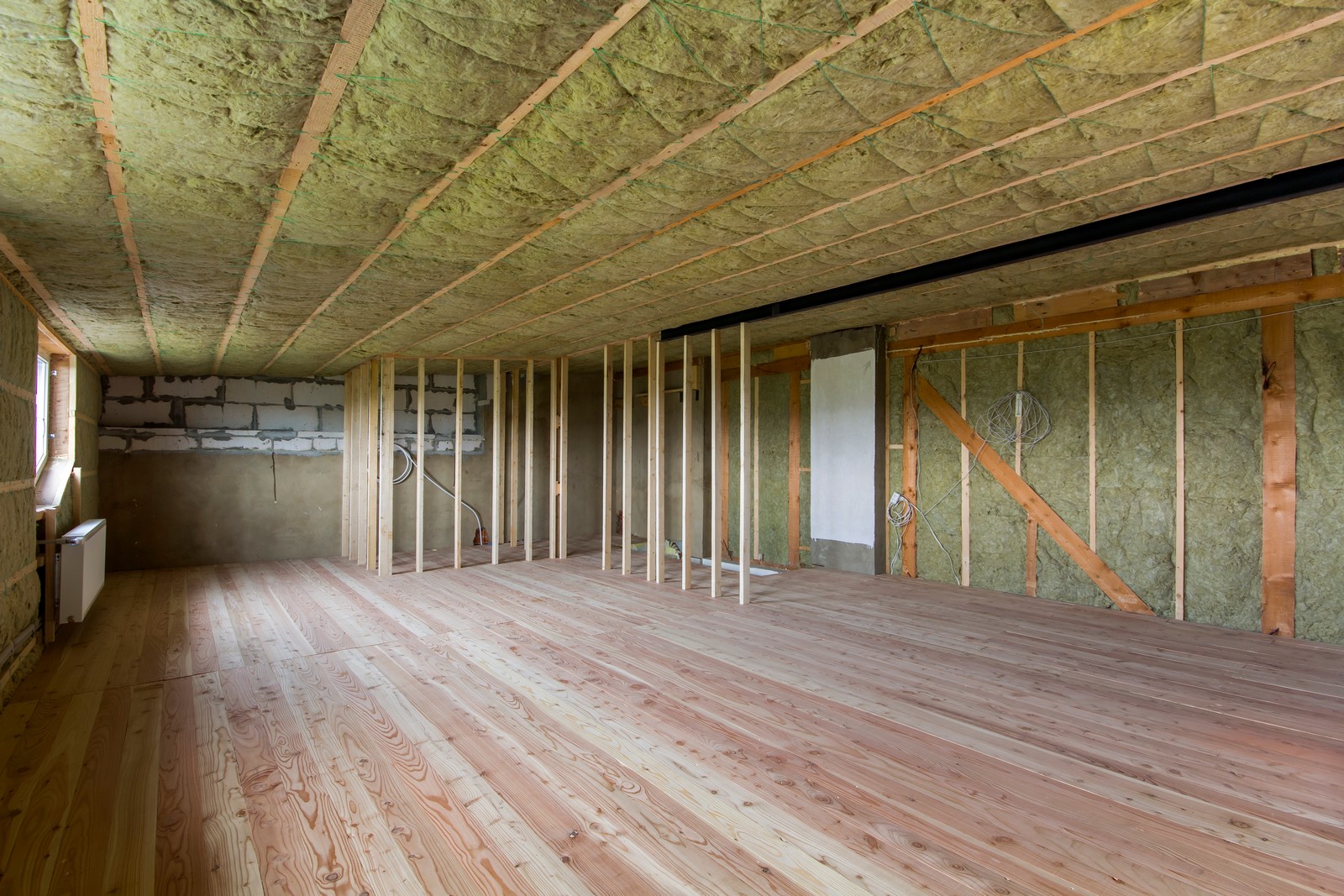 This image shows a room under construction or renovation. The floor appears to be newly laid with natural wood planking. The walls are partially insulated with fiberglass insulation, which is the yellowish material you see filling the gaps between the vertical wooden studs. The room is unfinished, with exposed wooden ceiling joists above and bare wooden wall studs visible. There are some wiring conduits or electrical boxes installed in the wall, and heating or plumbing pipes are present in the corner. The back wall features a section of concrete block that is partially coated with parging or plaster. It seems like this space may be part of a residential house, possibly in the attic or an upper floor, given the slope of the ceiling on the right side of the image.