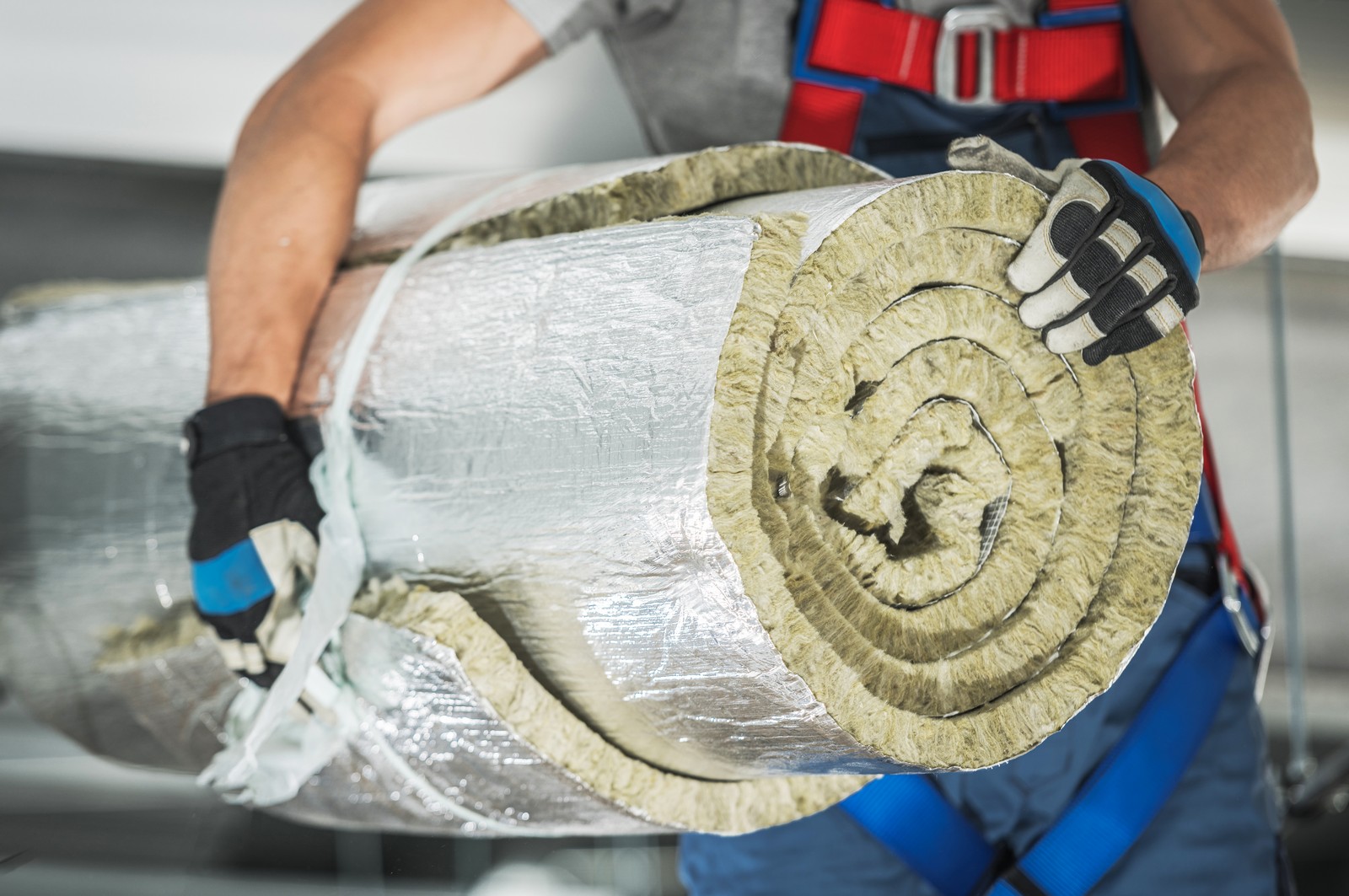 The image shows a person installing or handling a section of flexible ductwork, typically used in heating, ventilation, and air conditioning (HVAC) systems. The ductwork appears to be insulated and is wrapped in a reflective aluminium foil vapour barrier. The person is wearing work gloves and a tool belt, suggesting that they are a professional engaged in HVAC installation or maintenance activities. Only the person's torso and arms are visible, with a focus on the insulated duct being held.