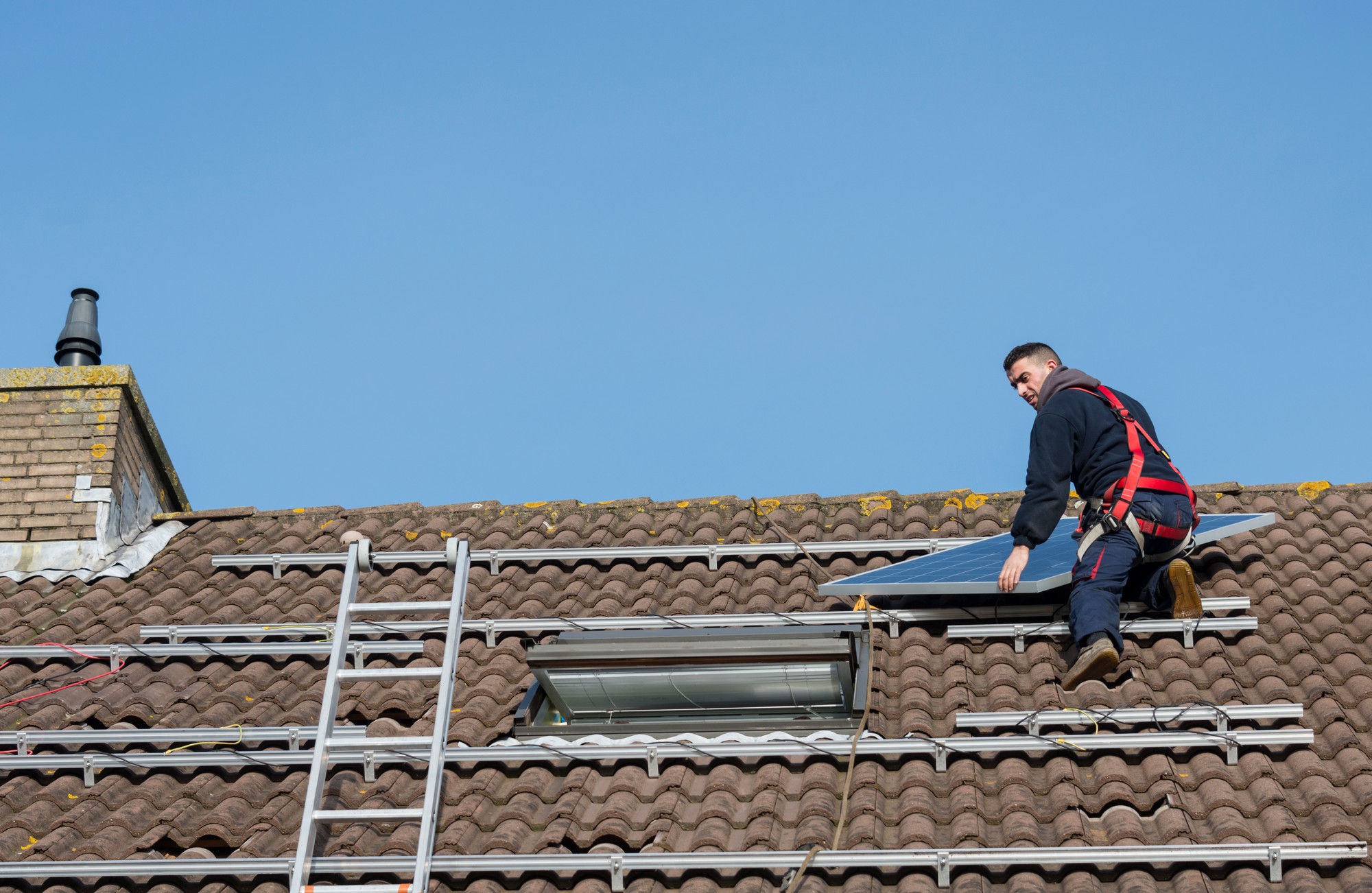 The image shows a person on a roof that is fitted with solar panels. The roof is covered with clay or concrete tiles, common in residential roofing. The person appears to be a worker, as they are wearing a safety harness, indicating that they might be installing, inspecting, or repairing the solar panels or the roof itself. They're also near a roof window, which suggests that the building is inhabited and may have living space underneath the roof. A ladder is placed against the side of the building, suggesting how the worker accessed the roof. The sky is clear, suggesting good weather conditions for outdoor work.