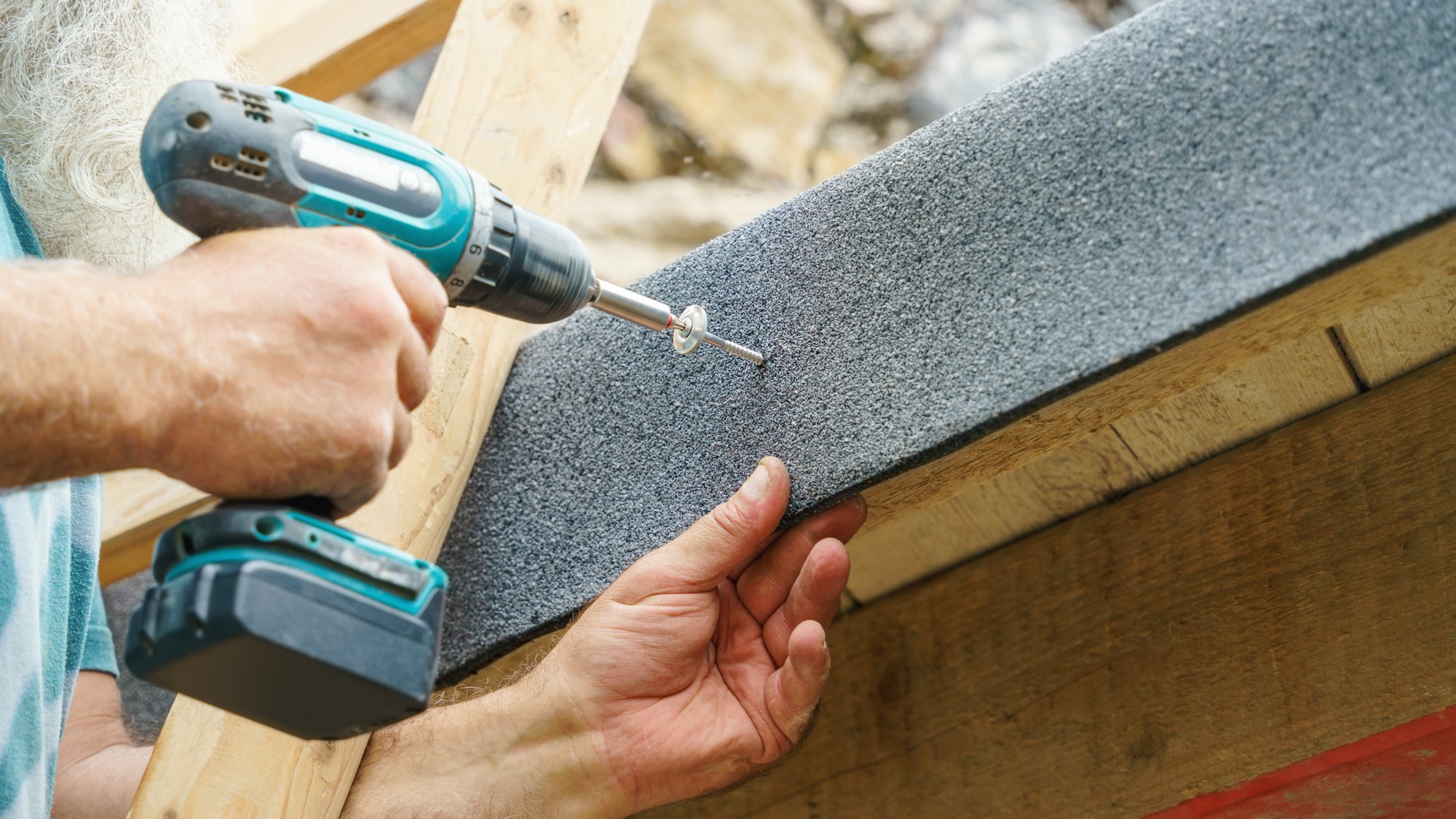 The image shows the hands of a person using a cordless power drill to drive a screw into a piece of non-slip, textured sheet material, which is being held against a wooden framework. It seems that this person is engaged in some sort of construction or DIY project where this material is being attached as a surface to the wood, possibly for traction or waterproofing purposes. The person is wearing a light blue shirt, and there are some stones and gravel in the background, indicating that the work might be taking place outdoor or in a workshop environment.