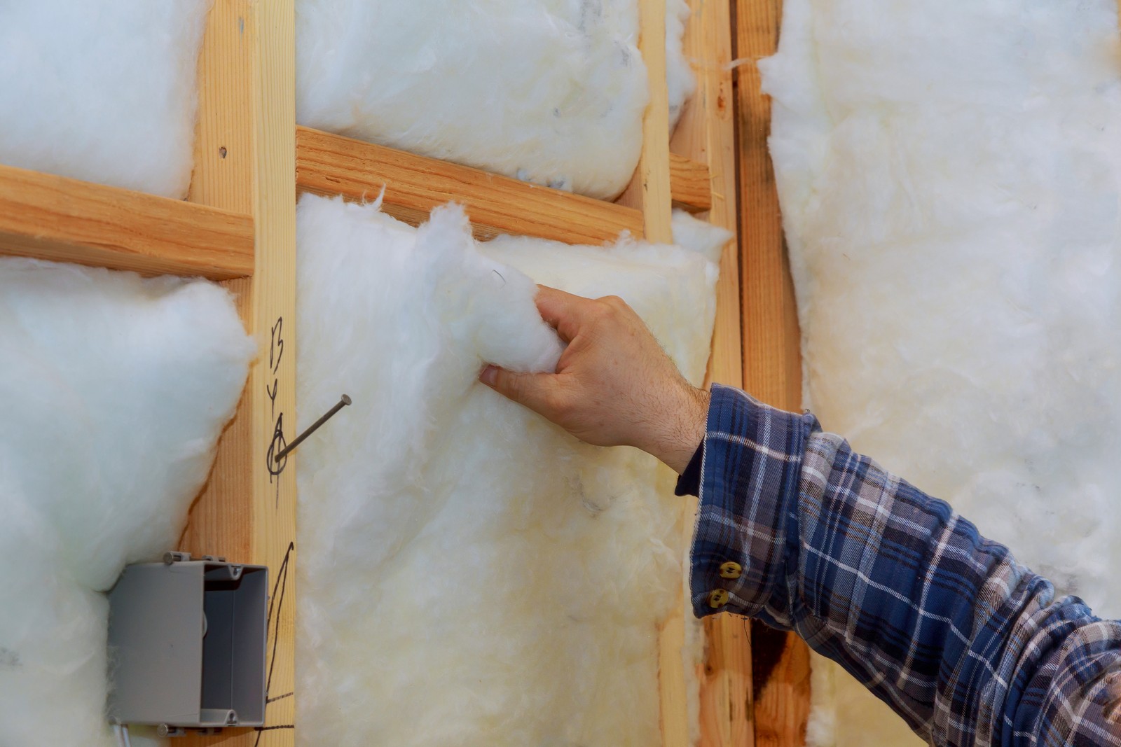 The image shows a person's arm and hand as they appear to be installing or inspecting insulation material in a wall frame made of wood. The wall studs are visible, and there is a gray electrical box mounted to one of them. The insulation material looks like it could be fiberglass, which is commonly used for thermal insulation in buildings to improve energy efficiency. The installation is not fully complete, as the insulation does not seem to be tightly fitted or sealed in some areas. The person is wearing a plaid shirt, suggesting they might be a construction worker, handyman, or homeowner engaged in a DIY project.