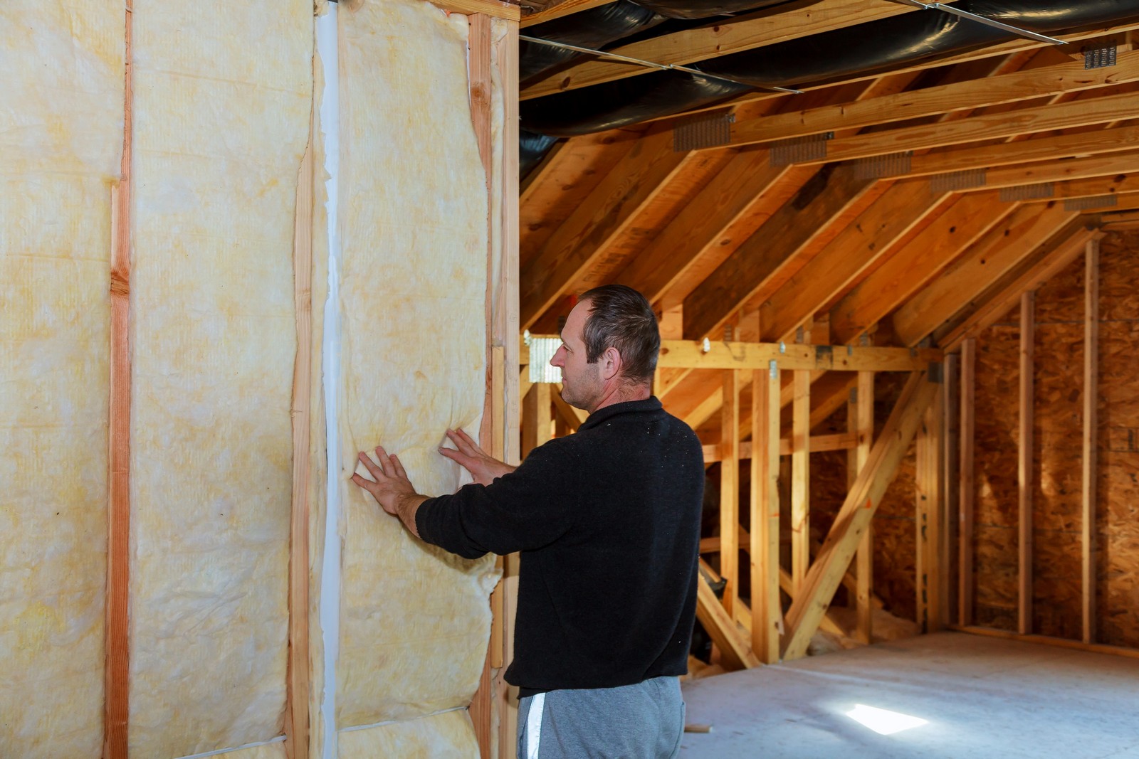 This image shows a construction or renovation scene where a man is installing insulation between the wooden studs of a wall in a building. The insulation appears to be fiberglass, which is commonly used to insulate buildings due to its thermal and acoustic properties. The environment looks like an unfinished attic space with visible roof trusses and sheathing, and you can see a vapour barrier on the ceiling, indicating ongoing work to make the space energy-efficient. The man is wearing casual clothing and seems to be engaged in do-it-yourself (DIY) or professional construction work.